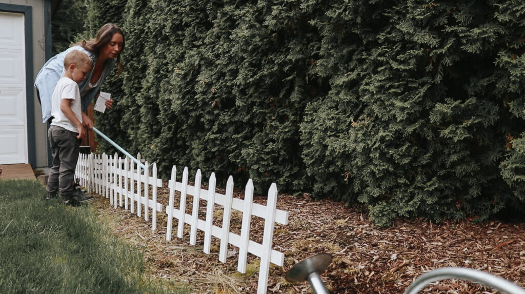 Christian homemaker waters garden with young son with a white picket fence in front of the garden.